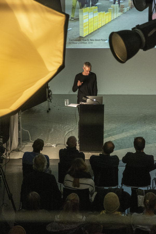 Birk Weiberg am Rednerpult mit Sicht auf Equipment des Fotostudios; Dozenten der Hochschule im Vordergrund