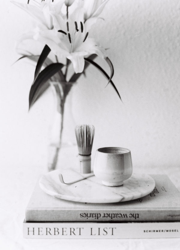 Matcha Besen und Tasse auf Tablett vor einer Blume in einer Vase.